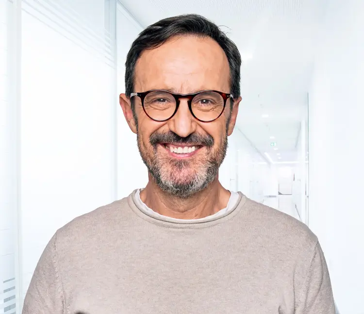 Porträt eines lächelnden Mannes mit Brille, Bart und hellem Pullover vor grauem Hintergrund