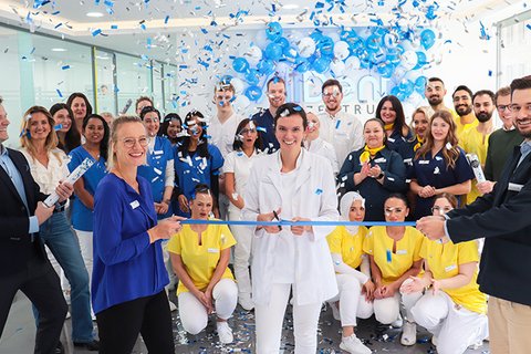 Menschen in bunter Arbeitskleidung schneiden ein blaues Band durch, während blaue Luftballons und Konfetti fallen.