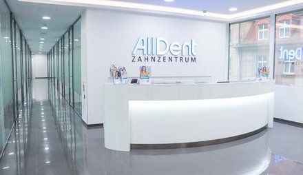 Empfangstresen in modernem Zahnarztzentrum mit beleuchtetem Schriftzug 'AllDent ZAHNZENTRUM' an weißer Wand und Glaswänden im Flur.