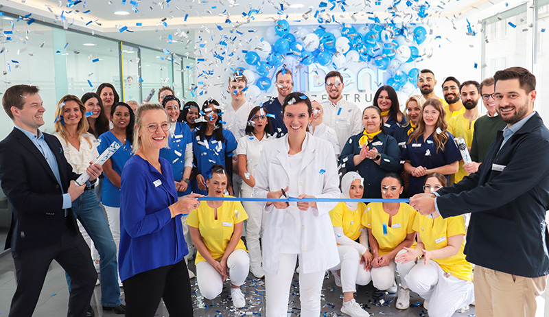 Menschen in bunter Arbeitskleidung schneiden ein blaues Band durch, während blaue Luftballons und Konfetti fallen.